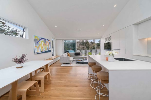 Bright, open-plan contemporary living and kitchen space with white walls and cabinetry, light timber flooring, a large white island with integrated sink, and stools. A dining table with timber chairs sits in the foreground, while a compact lounge area opens toward glazed doors and an outdoor view. Colourful wall art and understated furnishings add a playful accent.