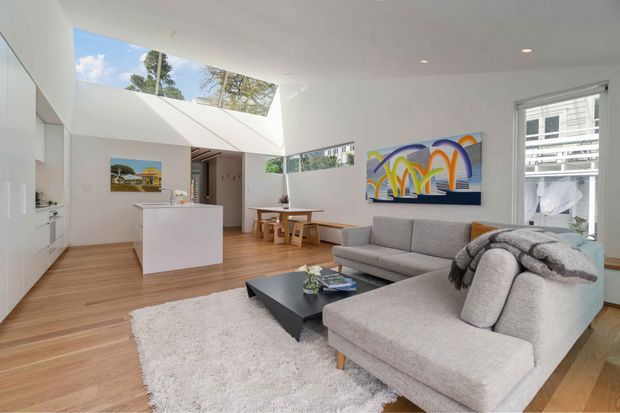 Bright open-plan contemporary living area with a kitchen at the rear, light timber flooring, white walls and cabinetry, a large grey sectional sofa, black coffee table, and white rug. A dramatic roof opening and skylight bring in natural light, with colourful artwork adding visual interest.