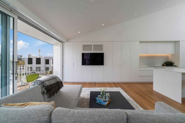 Bright contemporary open-plan living area with a large grey sectional sofa facing a black coffee table and a wall-mounted television integrated into extensive white built-in cabinetry. Warm timber flooring, recessed ceiling lights, and a minimalist white kitchen peninsula are visible, while full-height sliding glazing opens to a balcony with an urban view.