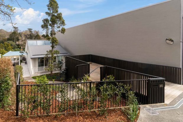 Exterior view of a contemporary building on a landscaped sloping site, featuring a large smooth light-grey rendered wall, black metal fencing and gates, timber decking, and a smaller pitched-roof structure with extensive glazing. The setting includes mature trees and a paved access area.