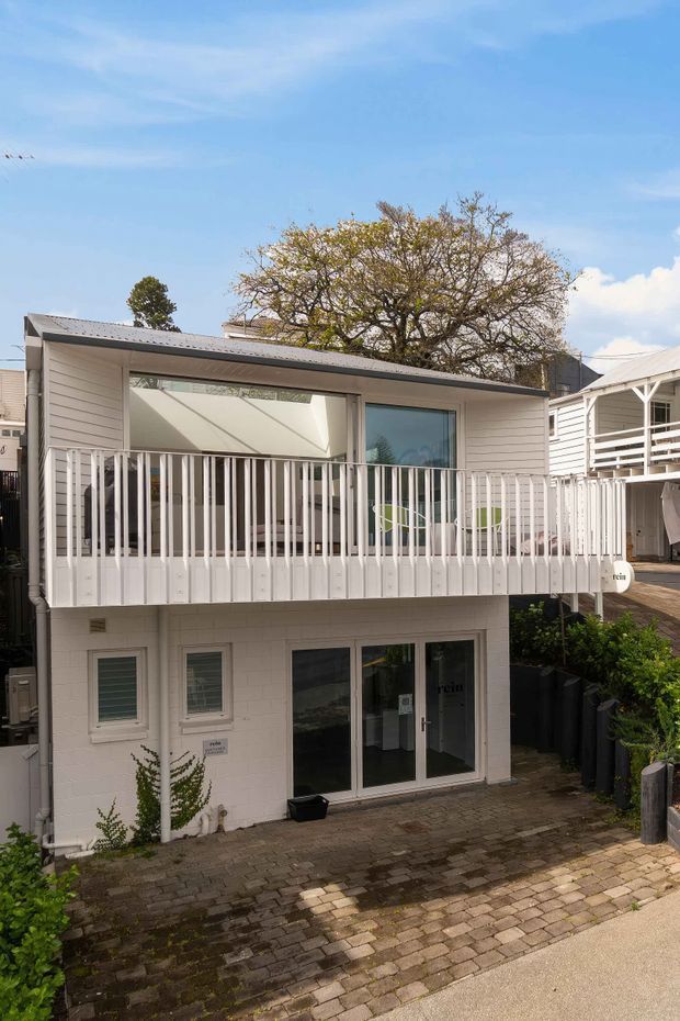 Exterior view of a compact two-storey white-clad building with a raised balcony, white vertical balustrade, large glazed openings, a low-pitched metal roof, and a paved forecourt. The building sits closely among neighbouring structures in an urban or suburban setting.