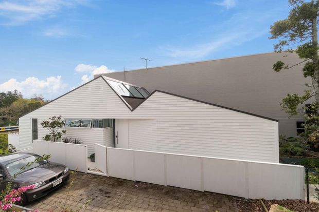 Exterior view of a contemporary residential building with overlapping pitched roof forms, light horizontal weatherboard cladding, dark roof surfaces, and a prominent glazed roof element. A paved driveway and privacy fencing occupy the foreground, with mature vegetation around the site.