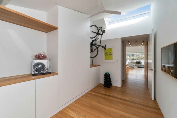 Bright contemporary hallway with built-in white cabinetry, warm timber shelving and timber flooring. Bicycles are mounted on the wall beside a small display niche, while a skylight and ceiling fan bring natural light into the circulation space. A living area is visible beyond the hallway, with a mirror reflecting part of the interior.