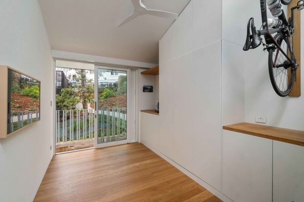 Compact, bright multipurpose room with warm timber flooring, white built-in cabinetry, open timber display shelves and a wall-mounted bicycle. Glazed sliding doors open to a small garden or courtyard, while a framed wall-mounted display is visible on the opposite wall.