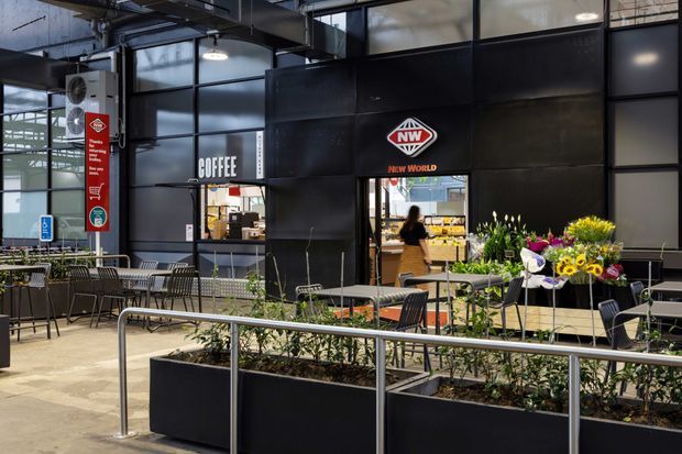 A modern indoor commercial retail and dining area featuring a black-panelled New World supermarket frontage, adjacent coffee counter, exposed services, large glazed windows, metal café tables and chairs, raised planter boxes, and stainless-steel accessibility railings.
