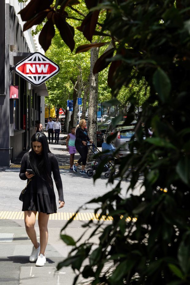 A leafy urban streetscape viewed along a pedestrian footpath, with a dark contemporary commercial building on the left, a prominent red-and-white NW directional sign, parked cars, street trees, pedestrians, and tactile paving at the crossing. Dense foreground foliage partially frames the scene.
