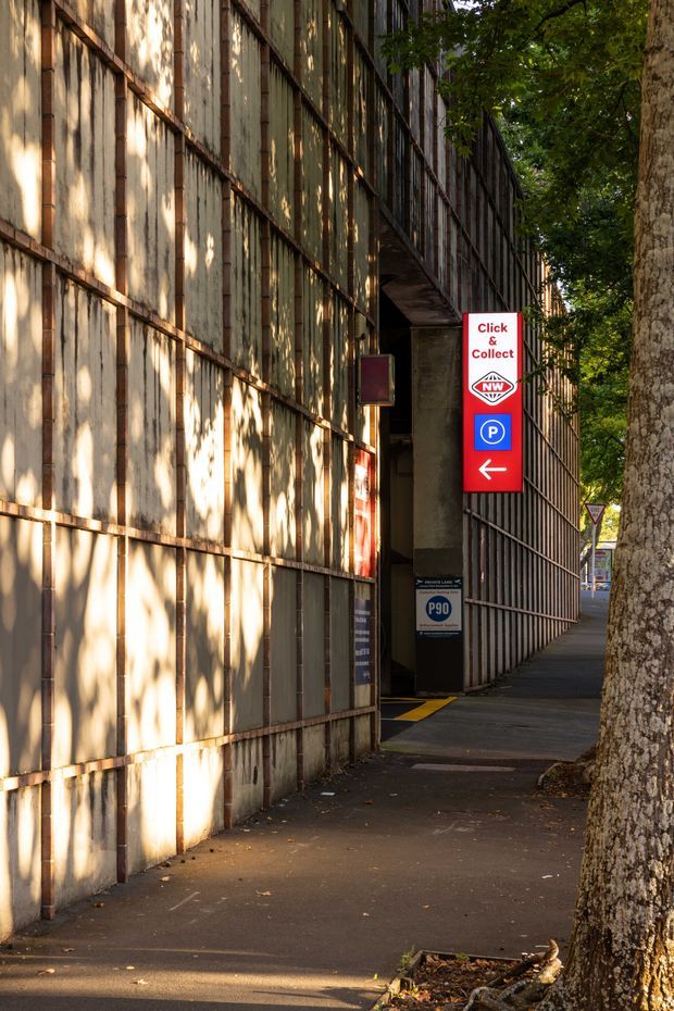 A narrow urban pedestrian footpath beside a tall, weathered metal-clad building. The facade is composed of large vertical and horizontal metal panels with a rusted, patinated finish, while a covered parking or collection entrance recedes into the building. Signage indicates click-and-collect parking and an arrow. Mature street trees line the sidewalk, with strong dappled sunlight and shadows across the facade and pavement.