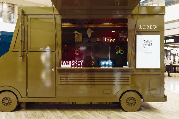 A vintage-style olive-gold mobile retail or food truck displayed inside a shopping mall. The vehicle has a service window with illuminated shelving, colourful decorative objects, neon-style words including “love,” “smile,” and “whiskey,” and a J.Crew branded sign. The polished mall floor and surrounding storefronts are visible in the background.