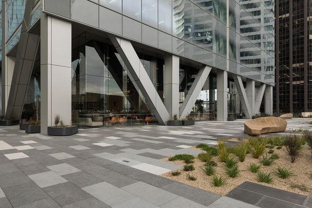 Contemporary commercial building frontage with a glass curtain wall, prominent white structural columns and diagonal bracing, and a broad paved plaza. The forecourt includes geometric grey pavers, low drought-tolerant planting in gravel beds, and sculptural timber seating. High-rise buildings are reflected in and visible through the glazing.
