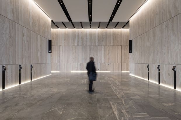 A large, symmetrical contemporary interior corridor or lobby clad in pale stone-look panels, with matching grey stone flooring, linear recessed lighting, dark ceiling tracks, and multiple elevator or door openings along the side walls. A person stands near the centre, emphasizing the generous scale and high ceiling.