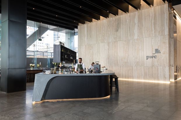 A refined commercial lobby or hospitality reception space featuring a sculptural dark bar or concierge counter, pale stone-clad feature wall with PwC branding, dark exposed ceiling beams, integrated linear lighting, and large glazed windows. The space has a polished, contemporary corporate aesthetic.