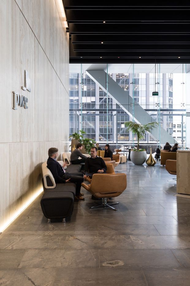 A sophisticated commercial lobby or reception lounge with a large pale timber-panelled feature wall bearing a raised logo, dark exposed linear ceiling battens, extensive floor-to-ceiling glazed curtain walling, and a dramatic diagonal structural element beyond. The space contains clustered tan and dark lounge chairs, small tables, planters, and people using the seating area. Large-format grey stone-look flooring and warm concealed wall lighting create a refined corporate atmosphere.