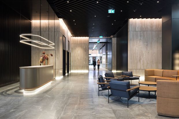 A sophisticated contemporary commercial lobby or reception area with a polished grey stone floor, dark ribbed ceiling and wall detailing, large pale stone wall panels, illuminated linear cove lighting, a sculptural ring pendant, and a curved reception desk. A lounge seating arrangement with upholstered armchairs, sofas, and low coffee tables occupies the foreground.