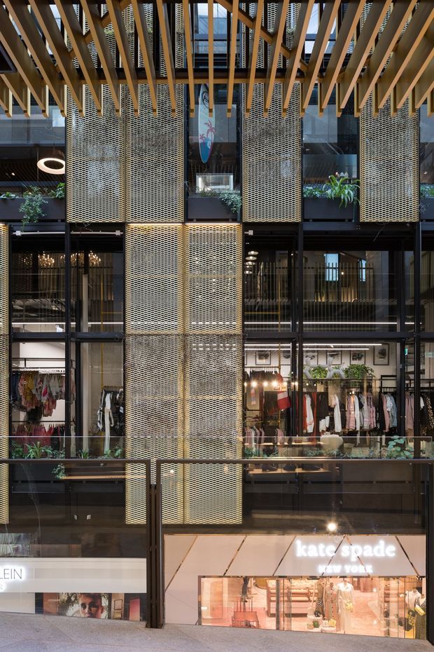 A multi-level contemporary retail interior or shopping arcade viewed across an open atrium. The facade is composed of black-framed glazing, warm brass-toned perforated metal screens, exposed timber slatted beams overhead, and planted balcony edges. Multiple fashion storefronts are visible, including Kate Spade, with pedestrian circulation in the foreground.