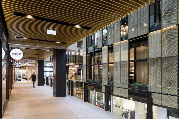 A contemporary multi-level indoor retail and commercial arcade with a wide tiled pedestrian walkway, shopfronts, dark structural columns, planted balcony edges, and a dramatic timber slatted ceiling. The facade on the right combines large glazed panels with perforated metal screens, while branded retail signage and lower-level storefronts are visible.