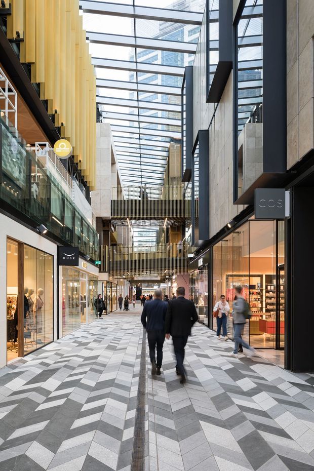 A contemporary multi-level indoor retail arcade or shopping centre with a dramatic glazed skylight roof, exposed structural steel, tall timber-toned vertical screens, concrete and stone walls, and glazed shopfronts lining a wide pedestrian walkway. The floor features a bold geometric chevron pattern in light and dark grey tiles. Shoppers move through the bright, modern commercial space.