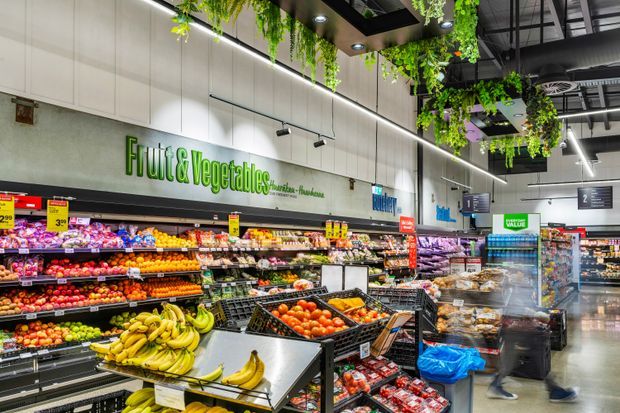 A brightly lit supermarket produce section with extensive displays of fresh fruit and vegetables. The space features high ceilings with exposed black services, suspended greenery, linear integrated lighting, and large illuminated "Fruit & Vegetables" signage.