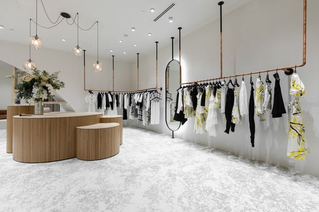 Bright, minimalist fashion retail interior with white walls, a pale patterned carpet, curved timber reception counter and stool, suspended pendant lights, exposed copper-toned clothing rails, full-length garments, and a freestanding arched mirror. The space has a clean, refined boutique atmosphere.