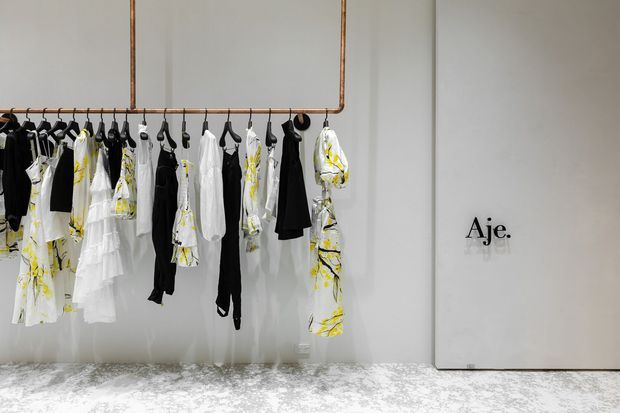 A minimalist fashion retail interior featuring a wall-mounted copper-toned clothing rail with black, white, and yellow garments hanging from it. A large pale panel with the “Aje.” logo stands to the right, while the space has smooth light walls and a mottled light grey floor.