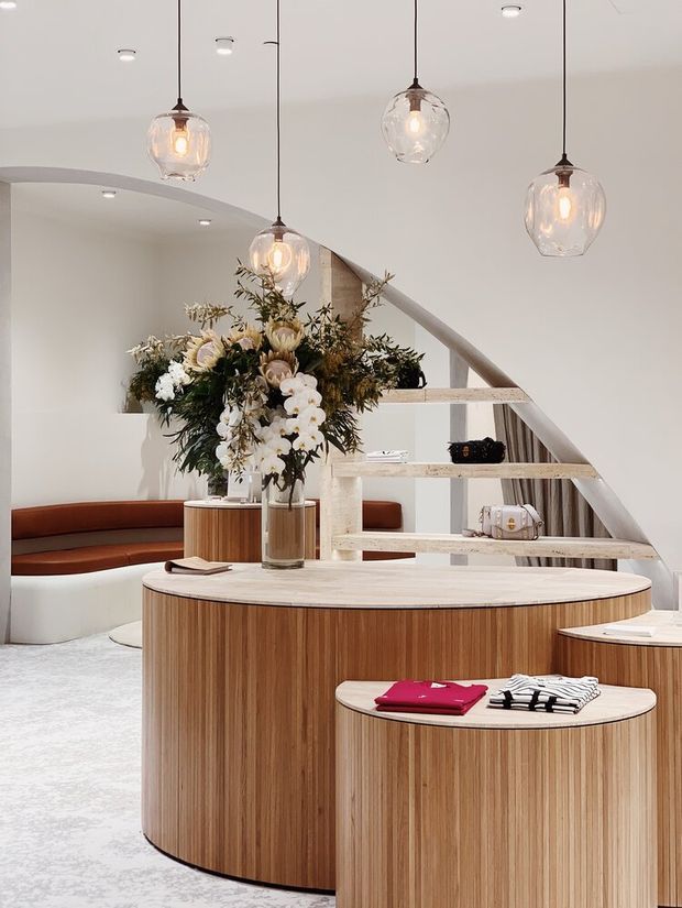 A refined retail or showroom interior with curved, vertically timber-clad display counters, integrated curved seating, and an open sculptural staircase. Multiple suspended clear-glass pendant lights illuminate a large floral arrangement and merchandise displays. The palette is predominantly warm timber, white, cream, and soft brown, with a polished contemporary fit-out.