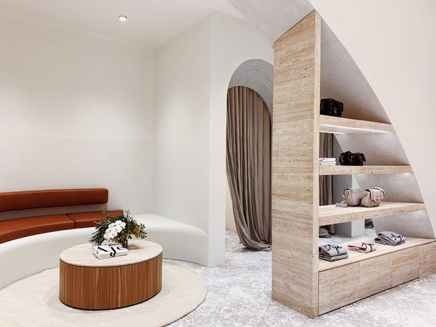 A refined contemporary retail or showroom interior featuring a curved built-in banquette in rust leather, a round ribbed timber coffee table, an off-white curved lounge seat, and a sculptural travertine-toned shelving partition with integrated display niches and drawers. An arched doorway with draped fabric leads to a secondary space. The palette is soft and neutral with warm timber and terracotta accents, and the flooring appears to be pale carpet or textured stone-like finish.