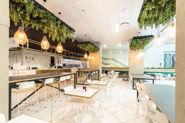 Bright contemporary commercial café or casual dining interior with extensive white tiled flooring, white tables and chairs, a long stone-topped counter with bar seating, and a compact service counter at the rear. Suspended warm pendant lights and abundant trailing greenery create a fresh, welcoming fit-out. A staircase and additional seating are visible toward the back, with large glazing on the right.