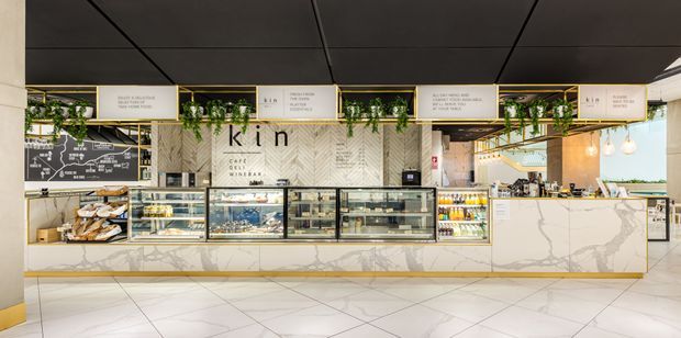 A contemporary café counter and food-service interior, viewed straight on. The long counter has white marble-look cladding with subtle grey veining and a slim brass-gold base trim, multiple glass display cases, refrigerated beverage shelving, and light timber shelving. Above, black ceiling panels and framed menu or branding boards are softened by trailing greenery. Pendant globe lights and pale geometric floor tiles complete the bright, refined commercial fit-out.
