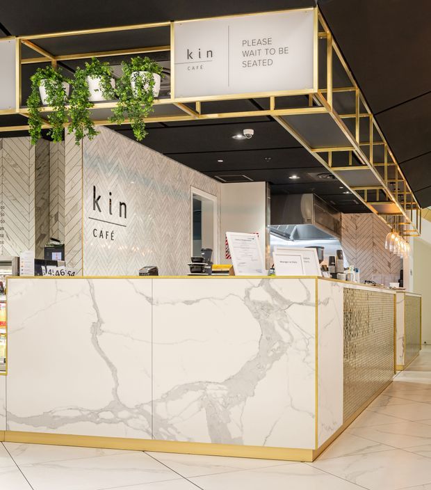 Interior view of a contemporary café counter and service area. The space features a large white marble-look counter with prominent grey veining, pale herringbone wall tiles, gold-toned metal framing and trim, black ceiling panels, hanging greenery, and a visible commercial kitchen behind the counter. Signage identifies the venue as Kin Café.