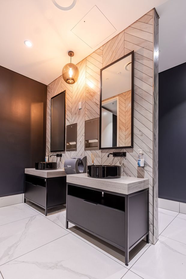 A stylish contemporary bathroom vanity area with two dark floating-style vanities, black vessel basins, pale stone-look benchtops, and large rectangular mirrors. The central feature wall is clad in pale herringbone-patterned timber or tile, contrasting with dark charcoal painted side walls. Large white marble-look floor tiles, black-framed mirrors, and warm pendant lighting create a refined, high-contrast finish.