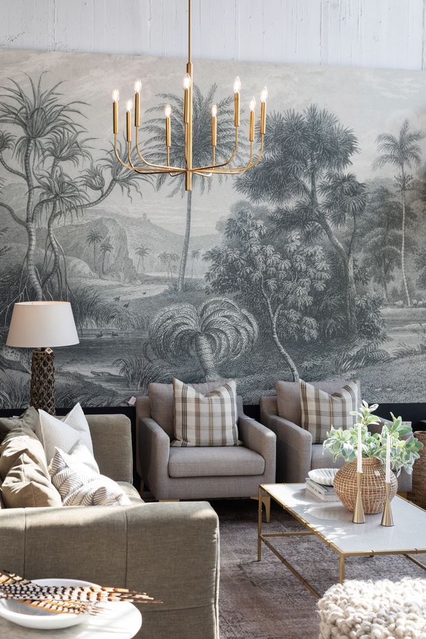 A refined living room with a large monochrome tropical landscape mural, grey upholstered seating, patterned cushions, a brass multi-arm chandelier, a sculptural floor lamp, and a white marble-topped coffee table with brass legs. Layered textiles and botanical accents create a sophisticated, warm interior.