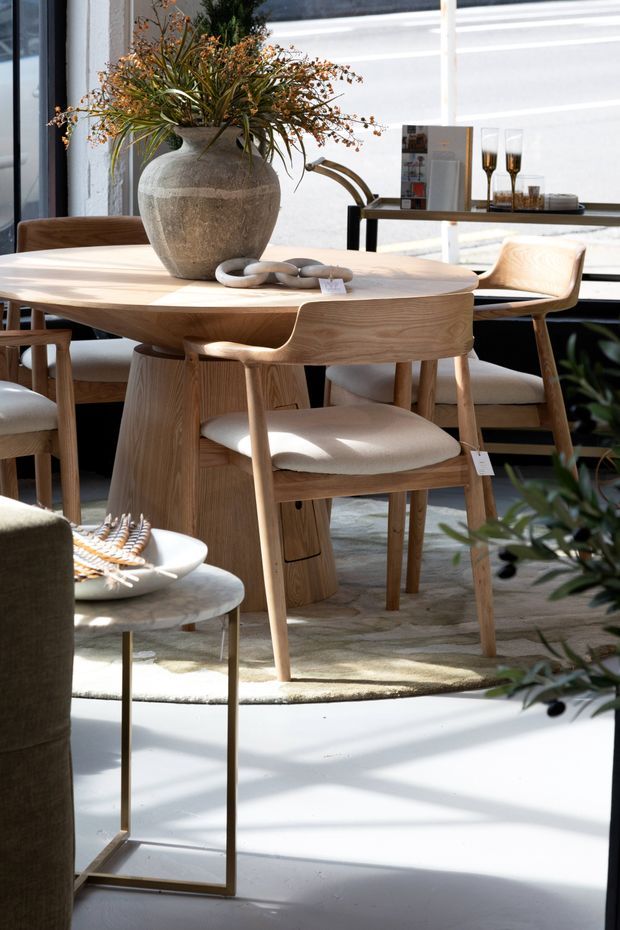 A bright, refined dining area featuring a round light-wood dining table with sculptural pedestal base, matching wooden dining chairs with upholstered cream seats, and a neutral rug over a pale floor. A large textured stone or ceramic vase with dried foliage is centred on the table. A slim black console with glassware sits behind the dining setting, while large black-framed windows bring in strong natural light.
