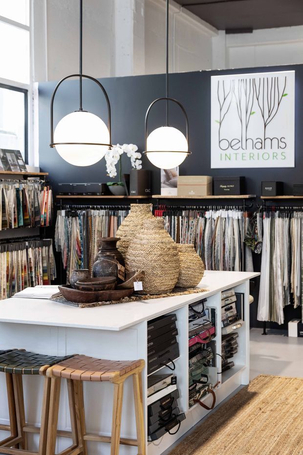 A stylish interior design and fabric showroom with floor-to-ceiling textile sample displays, a central white display island, woven stools, decorative pottery and baskets, and two globe pendant lights. The space has dark charcoal walls, white architectural surfaces, large windows, and a prominent Behams Interiors sign.