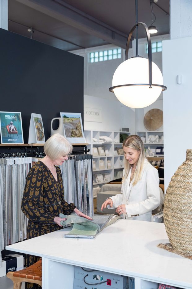 Bright, modern interior of a textile and homewares showroom. Two people stand behind a large white counter reviewing fabric samples, with fabric rolls and swatches displayed along the walls. The space features white painted walls, dark charcoal display cabinetry, exposed industrial-style ceiling elements, high-level glass block windows, open shelving, and a prominent spherical pendant light. A large woven basket or lampshade occupies the right foreground.