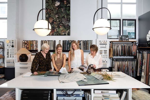 Bright, high-ceilinged interior of a textile or interior-furnishings showroom, with four people gathered around a large white display table reviewing fabric samples and design books. The room features extensive fabric sample racks, white built-in storage, large industrial-style windows, and two oversized globe pendant lights.