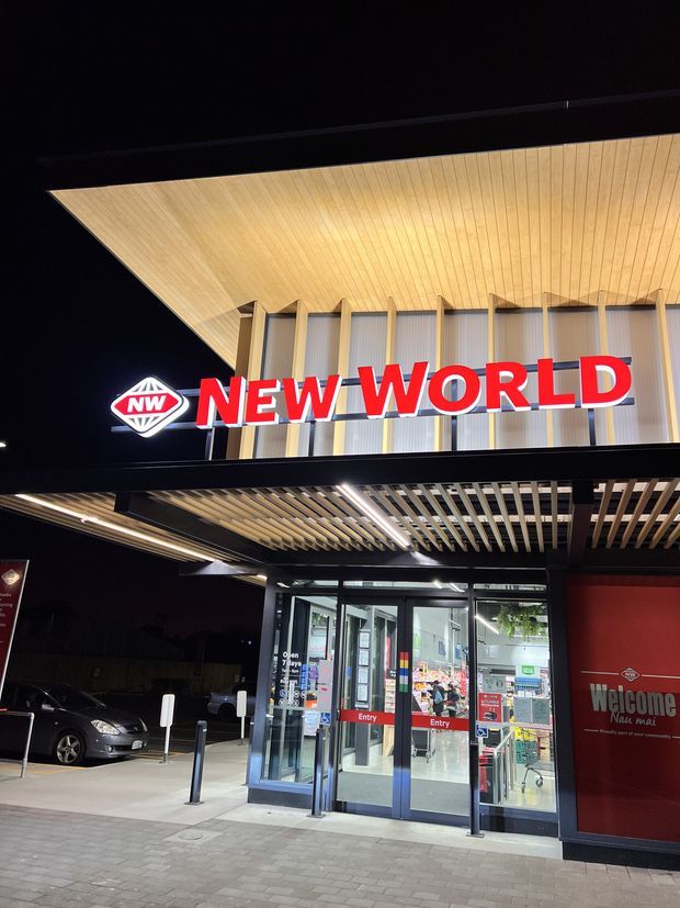 Night-time exterior photograph of a New World supermarket entrance. The commercial facade features a large illuminated red-and-white New World sign, a broad timber-toned soffit, vertical facade fins, dark framing, a slatted entrance canopy, automatic glass doors, and a red welcome panel.