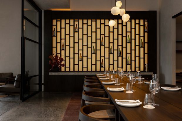 A moody contemporary dining space with a long dark timber dining table, upholstered bar-height chairs, and a sculptural cluster pendant light. The room is defined by a dramatic illuminated geometric partition or feature wall incorporating displayed bottles, with dark framed glass doors to the side and polished concrete flooring. Set tableware and glassware suggest a restaurant or hospitality interior.