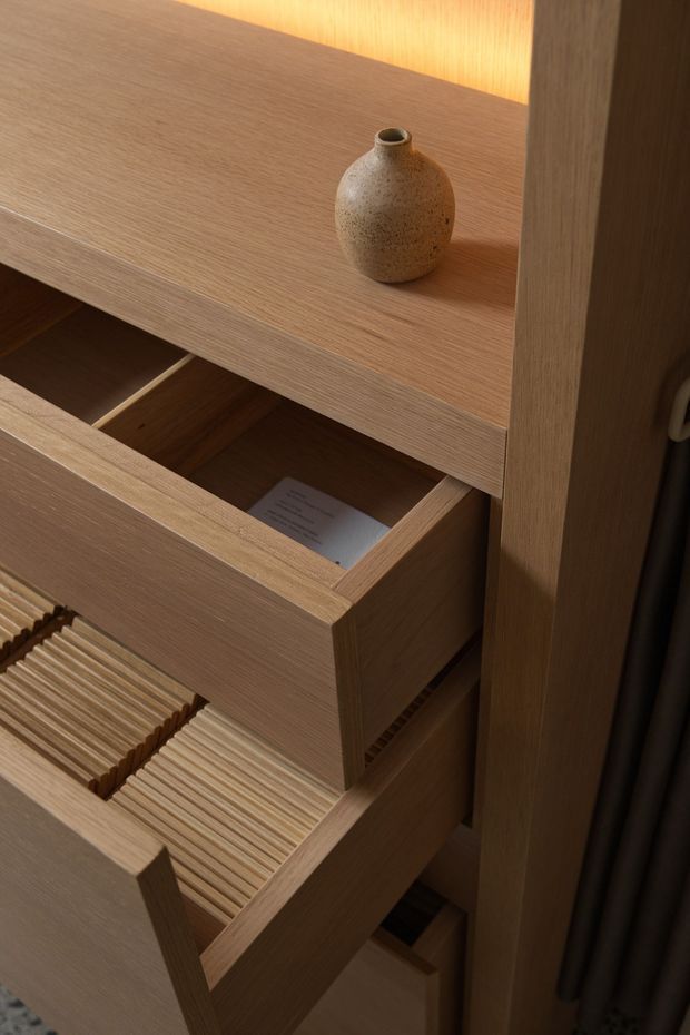 Close-up detail of a built-in timber storage unit with multiple open drawers, finely ribbed drawer organisers, and a small ceramic vase on the upper shelf. The warm wood grain and integrated warm lighting create a refined, bespoke joinery-focused composition.