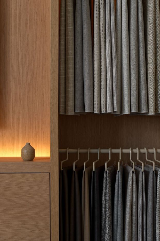 Close-up view of a refined walk-in wardrobe or dressing-room storage system, featuring warm wood-grain cabinetry, dark interior shelving, hanging garment samples, and neatly arranged fabric or clothing swatches. A softly lit display niche with a small textured ceramic vessel adds warmth and contrast.