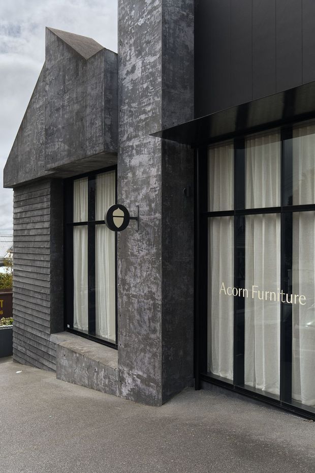 Exterior view of a dark, sculptural commercial storefront with large black-framed glazed windows, pale curtains, weathered grey concrete or cement-rendered walls, dark horizontal cladding, and a sharply angled projecting roof form. A round wall-mounted sign and the text “Acorn Furniture” identify the premises as a furniture showroom. The facade has a stark, monolithic contemporary appearance.
