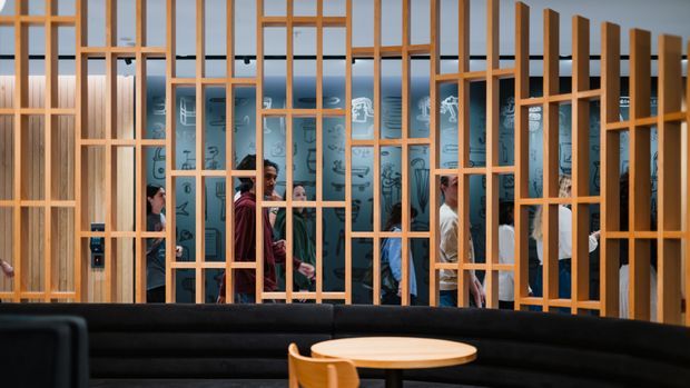 A contemporary commercial interior featuring a curved black upholstered banquette and round timber table in the foreground. A large open timber-framed screen divides the seating area from a busy communal space behind it, with blue illustrated wall graphics and several people visible beyond.