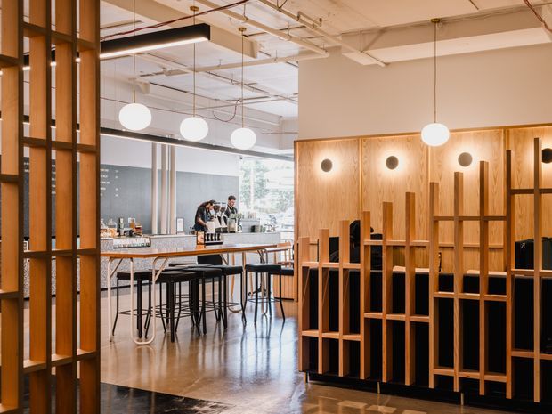 A contemporary commercial interior with an open café or hospitality counter, bar-height tables and stools, polished concrete flooring, and extensive warm timber screening and wall paneling. Suspended globe pendant lights, exposed ceiling services, black metal framing, and glazed partitions create a refined industrial atmosphere. The space appears to include a lounge or dining area beyond the timber partition.