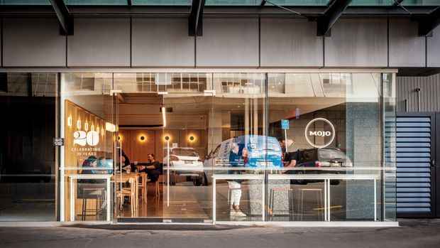 Street-facing view through full-height glazed storefront into a contemporary Mojo café. The commercial interior includes a service counter, coffee equipment, timber tables and chairs, warm pendant and wall lighting, and customers, framed by a dark metal and grey panelled building facade.