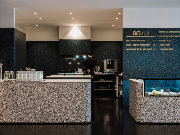 A dark, contemporary commercial food-service counter interior, likely a café or restaurant. The space features terrazzo-faced front counters, a glossy black floor, black textured wall finishes, integrated display refrigeration, menu lettering on the rear wall, recessed ceiling downlights, and commercial kitchen equipment behind the service counter.