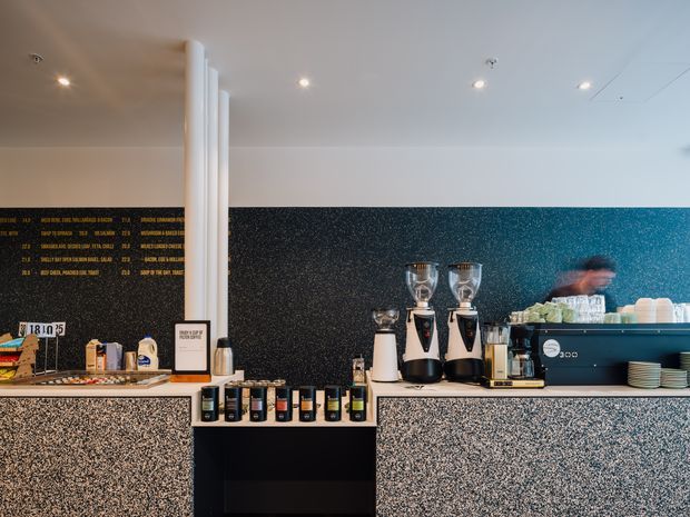 A contemporary café service counter with espresso machines, grinders, stacked cups, packaged goods, and a menu board. The counter and lower wall surfaces use a speckled terrazzo-like finish, while the dark textured backsplash creates strong contrast. White cylindrical structural columns and recessed ceiling lights contribute to a clean, modern commercial interior.