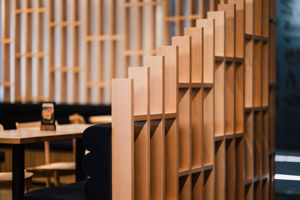 Interior view of a contemporary commercial or hospitality space featuring a prominent sculptural timber room divider with stepped vertical panels and integrated open shelving. Behind it is a dining or lounge area with tables, upholstered seating, and warm timber detailing. The space uses warm wood tones, black accents, and layered linear screens to create partial visual separation.