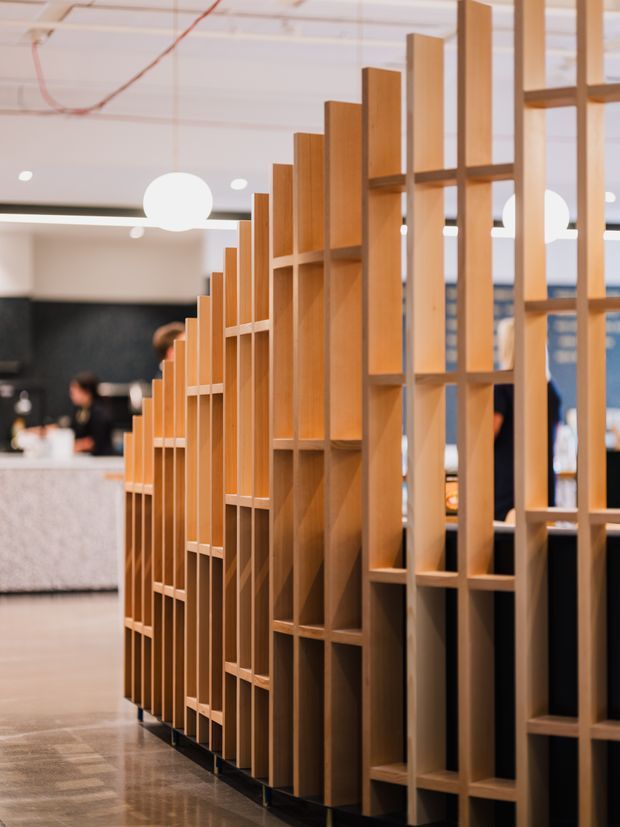 A contemporary commercial interior featuring a tall, rhythmic timber open-slat partition or shelving screen that divides the space while maintaining visual permeability. The warm wood structure has stepped vertical elements and integrated rectangular niches, set against a polished concrete floor, pale ceiling, globe pendant lights, and a blurred reception or hospitality area in the background.