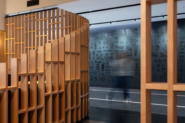 Interior view of a contemporary commercial or public space featuring a sculptural timber room divider and integrated open shelving in the foreground. Behind it is a dark illustrated feature wall with graphic line drawings, track lighting, and a polished dark floor. A blurred person is visible in the background, suggesting an active gallery, retail, or exhibition environment.