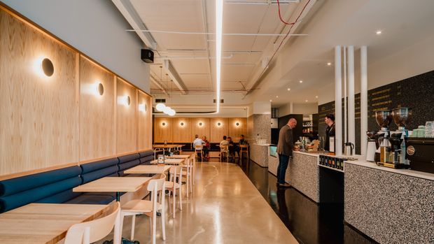 A warm, contemporary commercial café interior with a long blue upholstered banquette along a pale timber-panelled wall, small light-wood tables, and white chairs. The space has polished concrete flooring, a speckled stone service counter, exposed ceiling services, linear lighting, pendant lights, and a coffee preparation area. Customers and staff are visible in the background.