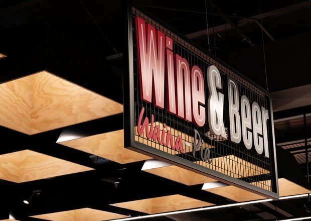A close-up view of a suspended, illuminated sign reading “Wine & Beef” with stylised lettering and smaller script text beneath. The sign hangs within a dark commercial interior, surrounded by exposed ceiling structure and multiple warm-toned plywood or timber ceiling light panels.