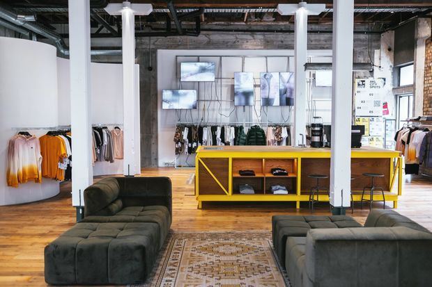 Industrial-style clothing retail showroom with exposed ceiling services, concrete walls, white structural columns, timber flooring, clothing racks, display screens, a bright yellow retail counter, and dark modular lounge seating around a patterned rug.